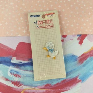 Puede incluir: Un paquete de "Fashionable Applique" de Wrights con un patito blanco con un lazo azul. La aplicación es para arte portátil. El paquete tiene un patrón de cuadrícula y una etiqueta de precio.