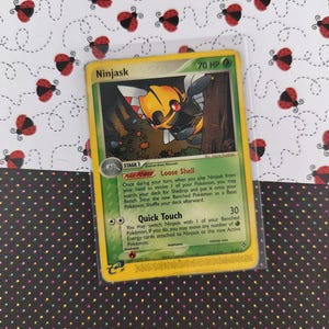 Pode incluir: Um cartão Pokémon Ninjask em uma manga protetora. O cartão apresenta uma criatura insetóide amarela e preta com detalhes em vermelho. O cartão tem uma borda verde e inclui texto descrevendo suas habilidades e HP.