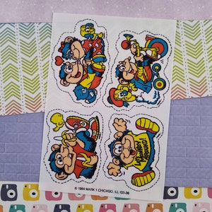 May include: Four vintage stickers featuring cartoon monkeys in various poses. The monkeys are wearing colourful clothing and are engaged in activities such as playing music, riding a tricycle, and eating a banana. The stickers are printed on white paper and have a cut-out design around the edges. The text on one sticker reads "Let's Monkey Around!"