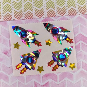 May include: Sheet of stickers featuring four holographic rocket ships with blue portholes and red and yellow flames, along with several gold star stickers. The stickers are on a white background, with a pink and green chevron pattern in the background.