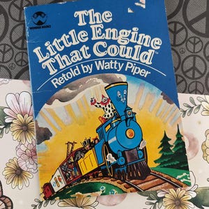 Könnte beinhalten: Ein Vintage-Exemplar von "The Little Engine That Could" von Watty Piper. Das Buchcover ist blau mit dem Titel in Weiß und einer farbenfrohen Illustration einer blauen Lokomotive mit einem lächelnden Gesicht, einem Clown und anderen Figuren.