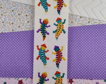 Pode incluir: Uma tira de autocolantes com desenhos de lagartos coloridos. Cada lagarto tem uma cabeça e um corpo geométricos com uma mistura de cores, incluindo vermelho, amarelo, azul e roxo. Os autocolantes estão sobre um fundo branco, rodeados por papel estampado.