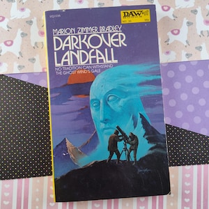 May include: A purple paperback book with a yellow border. The cover features a blue and white illustration of a face with a telescope in the foreground. The title is "Darkover Landfall" by Marion Zimmer Bradley. The book is a DAW book, number 36, and costs 95 pence.