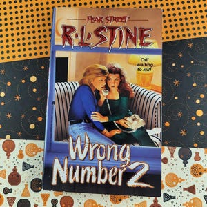 Puede incluir: Un libro de bolsillo vintage titulado "Wrong Number 2" de R.L. Stine de la serie Fear Street. La portada presenta a dos mujeres en un sofá a rayas, una de ellas sosteniendo un teléfono. También se ve el texto "Call waiting... to kill!".