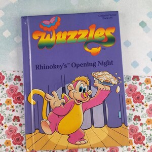 Könnte beinhalten: Ein lila "Wuzzles"-Buch, "Rhinokey's Opening Night", zeigt eine Zeichentrickfigur, die einen Kuchen hält. Der Titel ist in Regenbogenfarben gehalten. Das Buch gehört zur Collector Series, Buch Nr. 6.