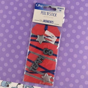 May include: A Peel 'N' Stick decorative tag with a red background and blue and silver stripes. The tag includes silver star and "GO" and "TEAM" embellishments. The package is labeled "My Moments".