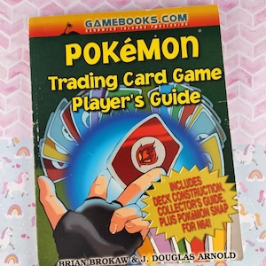 May include: A Pok&eacute;mon Trading Card Game Player's Guide book with a green cover and the title "POK&Eacute;MON" in large yellow letters. The cover features a hand holding cards and a yellow starburst with text. The book is by Brian Brokaw & J. Douglas Arnold.