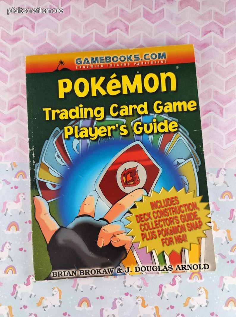 May include: A Pok&eacute;mon Trading Card Game Player's Guide book with a green cover and the title "POK&Eacute;MON" in large yellow letters. The cover features a hand holding cards and a yellow starburst with text. The book is by Brian Brokaw & J. Douglas Arnold.