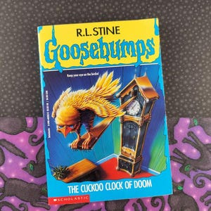 May include: A paperback book titled "Goosebumps: The Cuckoo Clock of Doom" by R.L. Stine. The cover features a yellow and blue design with a large bird and a grandfather clock. The book is from Scholastic.