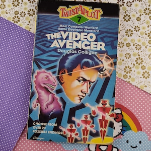 May include: A blue and red book cover with the title "The Video Avenger" and the author "Douglas Colligan". The book is part of the "Twistaplot" series and is number 7 in the series. The cover features a cartoon image of a man with a laser beam coming from his eyes and a pink dinosaur.