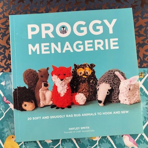 Könnte beinhalten: Ein Buch mit dem Titel "PROGGY MENAGERIE" mit türkisblauem Einband, das Rag Rug-Tierdesigns zeigt. Das Buch verspricht 20 weiche, kuschelige Tiere zum Häkeln und Nähen. Der Einband enthält auch den Namen der Autorin, Hayley Smith.