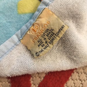 May include: A close-up of a white and blue cotton towel with a colorful pattern. A tag reads "FRANCO" and "ALL COTTON MADE IN BRAZIL &copy; THE WALT DISNEY COMPANY."