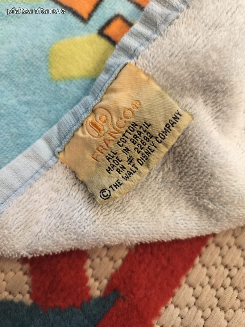 May include: A close-up of a white and blue cotton towel with a colorful pattern. A tag reads "FRANCO" and "ALL COTTON MADE IN BRAZIL &copy; THE WALT DISNEY COMPANY."