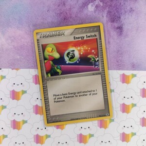 May include: A Pokémon "Energy Switch" trading card with a yellow border. The card features a green and red creature illustration with the text "Energy Switch" and instructions. The card is on a patterned background.