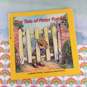 May include: A children's book titled "The Tale of Peter Rabbit" by Beatrix Potter, illustrated by David McPhail. The cover features an illustration of a rabbit in a blue jacket and red shoes, peeking through a white picket fence.