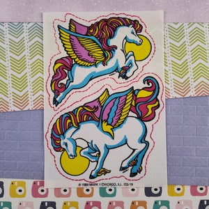 May include: Two vintage stickers featuring a white winged horse with a pink, yellow, and red mane and tail. The horse is depicted with wings outstretched and a yellow sun behind it. The stickers are on a white background with a red stitched border.
