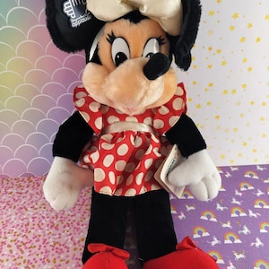 May include: A Minnie Mouse plush doll with black ears, a cream-colored bow, and a red and white polka dot dress. The doll has black pants, white gloves, and red shoes. The doll has a tag that says "applause".