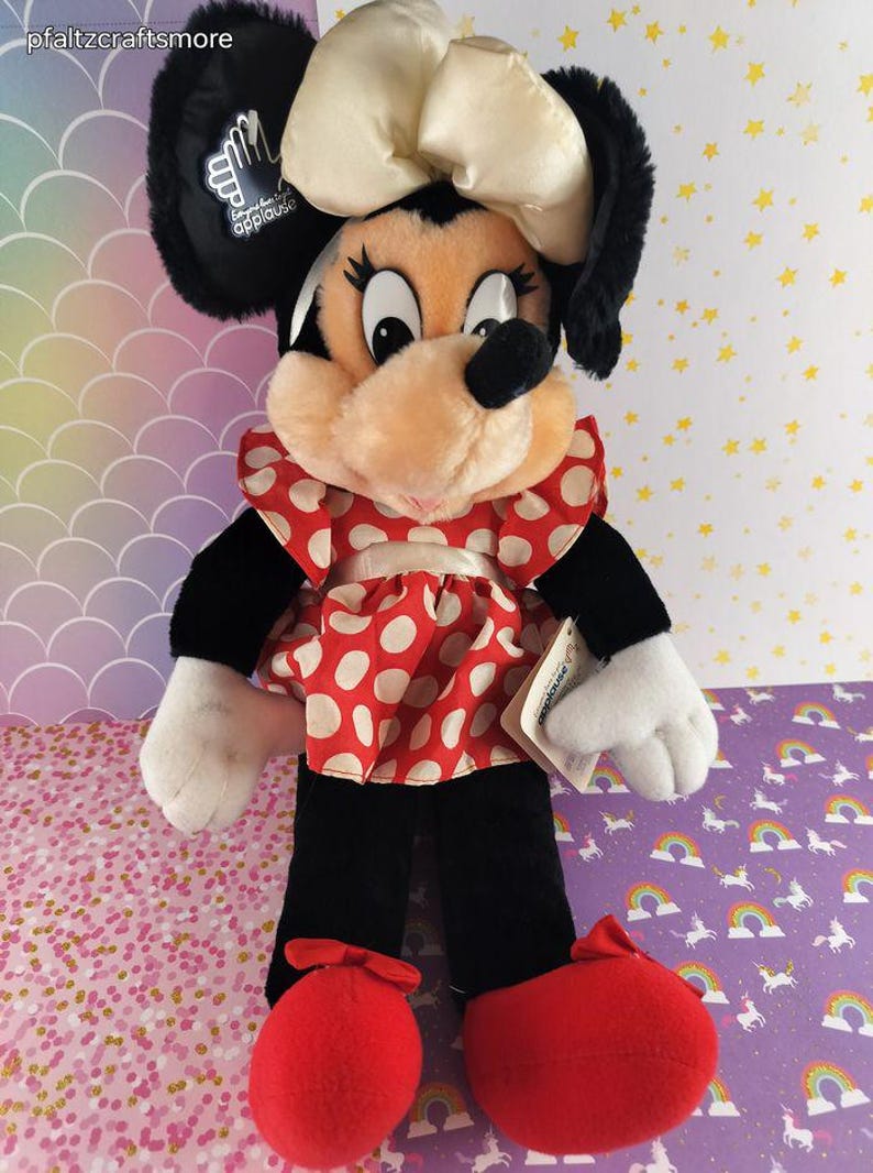 May include: A Minnie Mouse plush doll with black ears, a cream-colored bow, and a red and white polka dot dress. The doll has black pants, white gloves, and red shoes. The doll has a tag that says "applause".