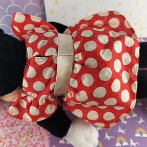 May include: A plush toy featuring a red dress with white polka dots and a matching bow. The bow is secured with a light beige ribbon. The toy has black arms and legs, and a white paw is visible.