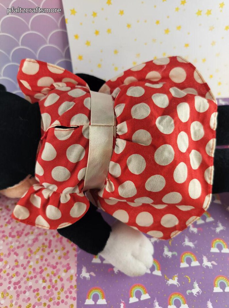 May include: A plush toy featuring a red dress with white polka dots and a matching bow. The bow is secured with a light beige ribbon. The toy has black arms and legs, and a white paw is visible.