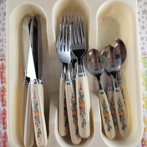 Op de afbeelding: Een vintage bestekset met crèmekleurige handvatten met een fruit- en bladerontwerp. De set bevat messen, vorken en lepels, netjes gerangschikt in een verdeeld dienblad. De handvatten hebben een zilveren band aan de basis.