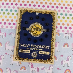 Puede incluir: Tarjeta vintage de broches de presión negros, talla 2-0, con el logotipo de la marca "Clinton". La tarjeta es azul marino con detalles dorados y el texto "Snap Fasteners Rustproof".