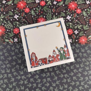 Peut inclure: Une carte carrée blanche avec une bordure bleue, ornée d'étoiles dorées et d'un croissant de lune. Le bas de la carte présente une ligne de personnages du Père Noël en robes rouges et vertes. La carte est posée sur un fond à motifs floraux.