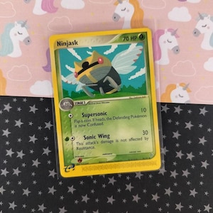 Pode incluir: Uma carta colecionável Pokémon Ninjask com borda amarela. A carta apresenta uma ilustração de uma criatura parecida com um inseto com asas, e o texto "Ninjask" e "70 HP". O fundo inclui um padrão de unicórnios cor-de-rosa.