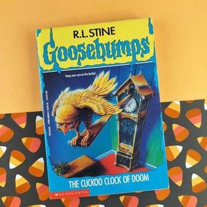May include: A paperback book titled "Goosebumps: The Cuckoo Clock of Doom" by R.L. Stine. The cover features a yellow bird with a clock and the text "Keep your eye on the birdie!" on a blue background. The book is on a patterned surface.