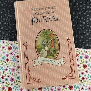 Può includere: Un diario rosa dell'edizione da collezione di Beatrix Potter. La copertina presenta strisce verticali, bordi decorativi e un'illustrazione ovale di Jemima Puddle-Duck con una volpe. Il titolo "JOURNAL" è stampato a grandi lettere nere.