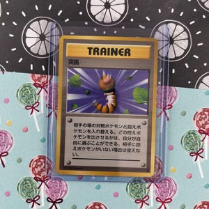 May include: A Trainer card featuring a cartoon creature with a brown and orange body, set against a purple background with green accents. The card is in a protective sleeve, with a background of lollipops and a black and white pattern.