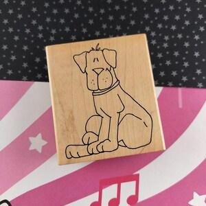 May include: A wooden stamp featuring a seated dog illustration. The dog is outlined in black, with a collar and a simple, friendly expression. The stamp is on a background of pink and white stripes and a black surface with white stars.
