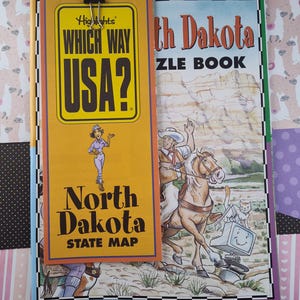 Puede incluir: Un libro amarillo y negro titulado "Which Way USA?" con un marcapáginas del mapa del estado de Dakota del Norte. El marcapáginas presenta a una niña de dibujos animados con una camisa morada y pantalones vaqueros azules. La portada del libro es verde y presenta a un vaquero de dibujos animados montando a caballo con un gato y una computadora.