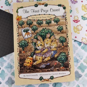 Puede incluir: Portada de libro infantil "The First-Prize Carrot", con ilustraciones de conejos en un jardín. Se muestran el título, el autor y el ilustrador. El fondo presenta papel estampado con personajes de dibujos animados.