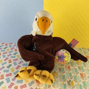 Puede incluir: Un águila de peluche marrón y blanca sosteniendo una botella verde y una bola de discoteca con una bandera estadounidense unida a su ala. El águila está sentada sobre una superficie estampada.