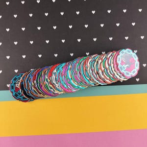 May include: A stack of colorful, circular craft pieces with various designs and colors. The pieces are arranged on a background of black paper with white heart shapes, and a strip of yellow, pink, and teal paper.