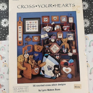 Può includere: Una copertina di libro intitolata "CROSS YOUR HEARTS" con disegni a punto croce, tra cui cuori, pezzi incorniciati e orsacchiotti. La copertina include anche il testo "16 counted cross stitch designs by Lynn Waters Busa."