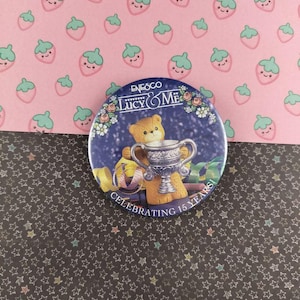 May include: A round pin with a teddy bear holding a trophy, with the text "Celebrating 15 Years!" and "Lucy & Me". The pin is set against a pink strawberry pattern and a black star pattern.