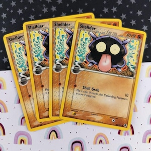 May include: Four Pokémon trading cards featuring the character Shellder. The cards have a yellow border and a brown background with the Shellder illustration. The text on the cards includes "Shell Grab" and "Flip a coin. If heads, the Defending Pokémon is now Paralyzed."