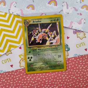 May include: A Pokemon trading card featuring Ariados, a bug-type Pokemon with a yellow and orange body, long legs, and a red stinger. The card has a 60 HP value and the text "Spider Web" and "Poison Bite".