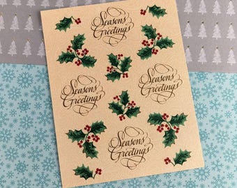 May include: A sheet of cream-colored stickers with the words "Seasons Greetings" in elegant script, along with illustrations of holly leaves and red berries. The stickers are on a background of snowflakes and Christmas trees.