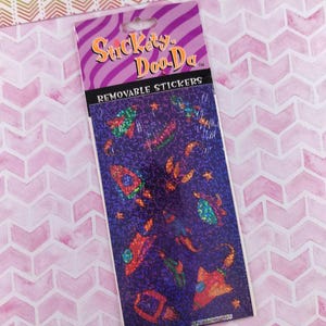 May include: A package of "Suckers-Doo-Da" removable stickers. The stickers feature a space theme with rockets, planets, and stars in vibrant colors. The background is a dark purple with a holographic effect.