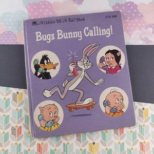 May include: A vintage "Bugs Bunny Calling!" Golden Tell-A-Tale Book. The cover is purple with Bugs Bunny on a skateboard, holding a phone. Other Looney Tunes characters are also featured. The book has a U.S. price of 69¢.