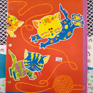 Puede incluir: Una libreta roja con un borde azul y morado. La portada presenta tres gatos de dibujos animados coloridos jugando con una bola de lana. El texto "Rite Aid 83¢" está en la portada.