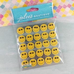 Könnte beinhalten: Eine Packung mit 25 gelben Smiley-Aufklebern mit schwarzen Augen und Mund. Die runden Aufkleber sind glitzernd und eignen sich für Bastelarbeiten. Auf der Verpackung steht "jolee's BOUTIQUE".