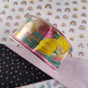 May include: A roll of pink and green ribbon with a floral and lemon print. The ribbon is on a white background with a pattern of small rainbows.