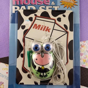 Könnte beinhalten: Eine grüne und weiße, kuhförmige Computermaus mit großen blauen Augen und ein Milchkarton-förmiges Mauspad mit schwarz-weißem Kuhmuster. Die Verpackung trägt die Aufschrift "mouse & pad set" und "fun animal designs!"