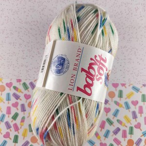 May include: A skein of Lion Brand Baby Soft yarn in a cream color with colorful stripes. The label reads "Baby Soft" in pink script. The yarn is on a patterned background of ice pops.