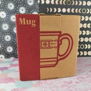 Puede incluir: Una caja de cartón con un lado rojo con la etiqueta "Mug". La caja presenta un contorno rojo de un diseño de taza. El fondo incluye papel estampado con diseños en blanco y negro y patrones en rosa y blanco.