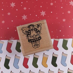 May include: A wooden stamp featuring a whimsical angel design with plaid accents. The stamp is set against a red background with white snowflakes and a patterned paper with Christmas stockings. Ideal for crafting and holiday projects.